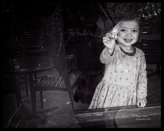 A young girl in a patterned dress smiles while holding an object up to a window. The scene is black and white, with reflections of tables and other people visible in the background.