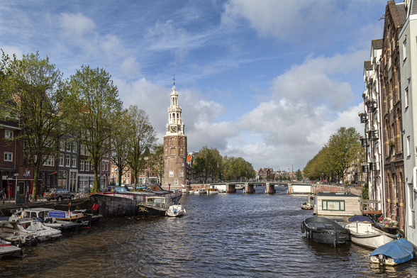 It's a bright day with some patchy, light clouds in the blue sky. We're in a city and on a low pedestrian bridge crossing over a wide canal. Small boats are tied up along both sides with those on the right side directly up against the walls of tall town houses lining that bank. On the left bank there's a small rise to a narrow road in front of buildings with shops on the ground floor and accommodation on the three or four floors above. A few cars and bikes can be seen parked up and there are some trees spaced apart in front, taller than the buildings but sparse with leaves. Further up the canal there's another gently arched bridge crossing it with just enough space beneath its three gaps in the span to allow rowboats or similarly-sized vessels through. An octagonal brick tower that might have been part of a church or council building with white upper clock mounting is to the left of the bridge and beyond it we can see more trees and buildings as the canal disappears into the distance.