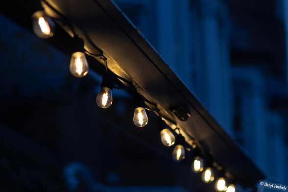 A string of lit bulbs hanging from a beam, at dusk. 