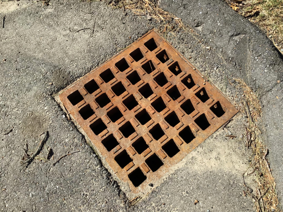 A closeup of a square rusted drainage grate in bright sunlight with square holes and little protuberances between the holes.