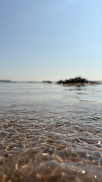 Gentle ripples of water wash over a sandy shore with a distant view of rocks and a clear blue sky.