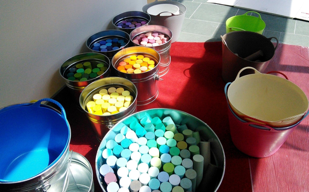 Several tinned buckets holding brightly-painted wooden dowels and some colored plastic buckets, in bright sunlight but with shadows falling across parts.
