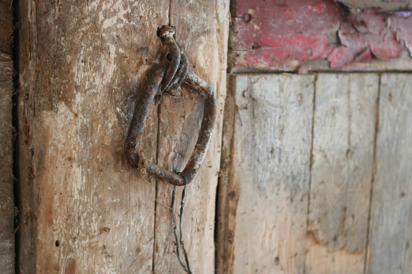 Close up photo of a metal square ring fixed to a wall of wood planks by two folded nails. The ring and the nails are rusted, have old paint and dust on them, and there is a plastic black tie wrap hanging on the ring. The vertical wood planks are brown and grey with a whitewashed colour and one horizontal plank on the top right is a window sill and has flaky red paint.