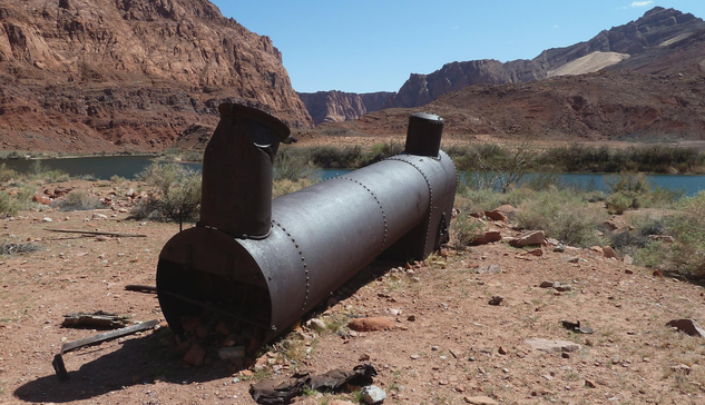 Photograph of an abandoned boiler, rusted and now dark brown with a deep red hue. The boiler sits on the edge of a river, with blue water and green and grey bushes on its banks, in a red desert landscape typical of southwest US, with mesas and canyons on each side of the river.