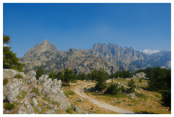 Panorama Photo of a mountain range in the distance. Blue sky. A path and green trees in the foreground.