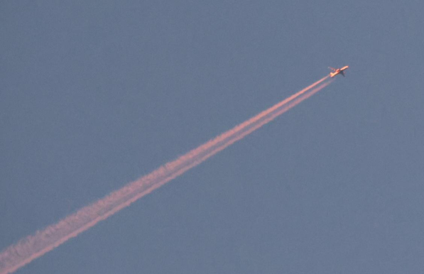 Ein Flugzeug mit jeweils einem Düsentriebwerke an den Tragflächen, leuchtet rot - der Sonnenaufgang ist erst in 35 Minuten vorhergesagt. Des Weiteren sieht man am blauen Himmel keine Wolken  nur den Abgasstrahl des Flugzeuges, welcher ebenfalls in der Farbe rot erstrahlt. 
Matzework 