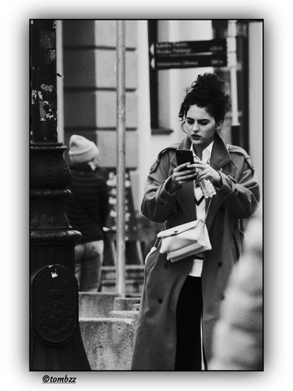 A young woman is walking down the street, eyes fixed on the screen of a smartphone held in both hands. She has dark, curly hair styled in a loose bun. She’s wearing a long, light-colored coat with large buttons, under which a striped sweater and a light shirt are visible. On her shoulder, she carries a light handbag with a chain strap. On the left side of the frame, part of a streetlamp is visible. In the background, the blurred figure of another woman and elements of urban architecture. The entire scene is captured in the black-and-white aesthetic of street photography.