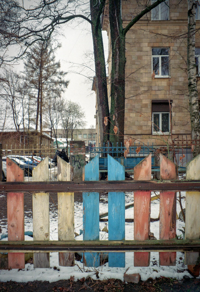 The photo shows a multi-colored fence, behind which a building and a tree are visible. There is snow under the fence.