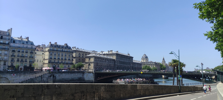 Vue urbaine lumineuse à Paris, prise depuis les quais de Seine. On aperçoit un pont noir élégant, des bâtiments haussmanniens baignés de soleil, une péniche remplie de touristes, et un ciel bleu éclatant sans nuage. Deux promeneurs descendent le quai sur la droite, encadrés par des palmiers urbains et une perspective calme. L’ambiance est claire, posée, doucement vivante