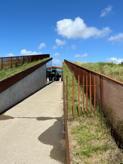 Ein Weg aus Beton in ein Gebäude, welches sich aus der Landschaft mit begrünten Dächern heraushebt. Die Dächer sind begehbar und haben dadurch umlaufende Absturzsicherungen aus Cortenstahl.
