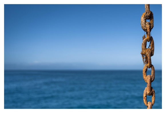 The picture shows a scene by the sea. A rusty chain can be seen in the foreground, which runs vertically and is presumably attached to a ship or a mooring. The chain has a reddish-brown color due to rust and consists of large, oval links. A calm, deep blue sea stretches out in the background, merging with a clear, light blue sky. The horizon line is gentle and gives the picture a peaceful atmosphere. There are no other noticeable details or objects in the picture.