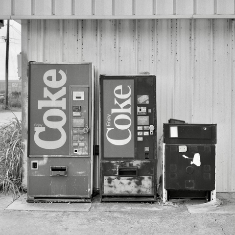 A black and white film photo. Two old soda vending machines sit side-by-side with an old, discarded stove in Taylor, Texas. Sunday, June 15, 2025.