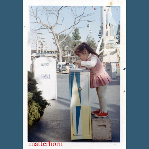 A young girl stands on a concrete block, reaching into a colorful, vertically striped box in an amusement park setting. She is wearing a pink dress and white leggings, with red shoes. Background features amusement park attractions and trees.