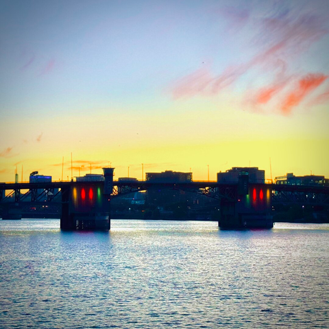 The Morrison Bridge lights up in red, yellow, and green colors over the Willamette River just before sunrise under a yellow, blue, and pink-cloudy sky.