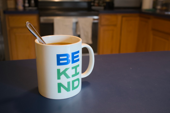 A mug of tea on a counter in a kitchen. There’s a spoon sitting in the mug and the mug reads BE KIND.