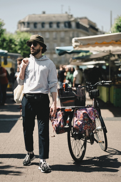 A bearded man wearing a bucket hat, sunglasses, and a light hoodie stands beside a bicycle with colorful floral saddle bags at an outdoor market. He carries a tote bag over his shoulder, with a relaxed posture, surrounded by people and market stalls under canopies on a sunny day.