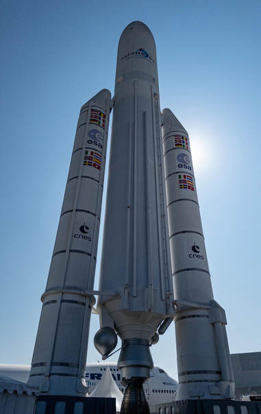 A full-size model of the giant Ariane 5 rocket, with its core stage and twin boosters. The rocket is white with flags and logos from ESA, CNES, and the member states in the Ariane programme. The sun is behind the rocket.