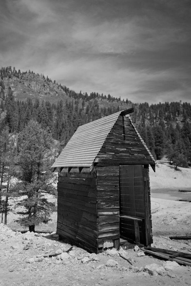 Black and white photo of a small, weathered wooden shack with a corrugated metal roof, standing alone in a rocky clearing with forested hills behind it at Sulphur Springs near Valles Caldera National Preserve.