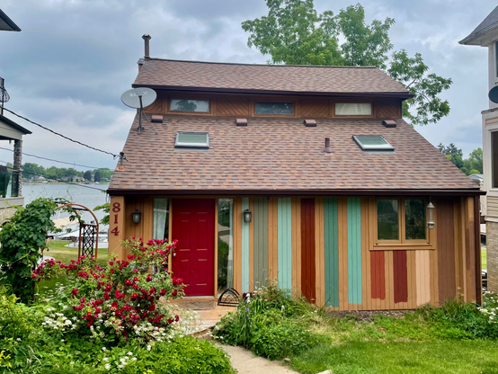 Photograph of a small two story brown house with stripes of various colors in the background. There is a lake in the background. The house is flanked on either side by much larger houses that tower over it, but hey, we’re mostly looking out at the lake not at the walls of our neighbors’ houses. Also, please take a moment to appreciate the spectacular rose bush in the front of the red front door to the left of the house. The rose bush is a carpet of red roses surrounding by white flowers from the ground cover. It’s overgrown and my wife frets that it’s a mess, but, man, that is some rose bush!