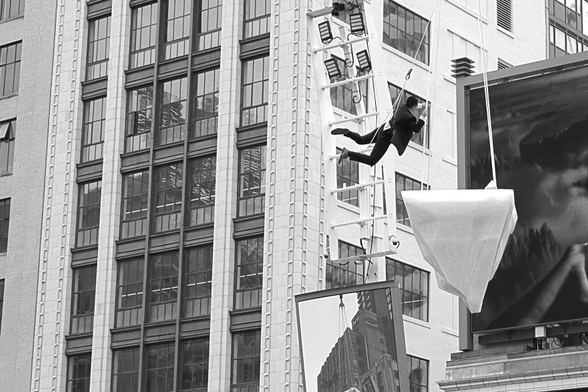 Suspended over the square, a performer seems to be flying in mid air above a suspended platform as part of a performance art piece. Behind him at the right edge of the frame is the edge of a huge LCD billboard. In the left background is the white facade of a downtown building filling the frame from top to bottom.
