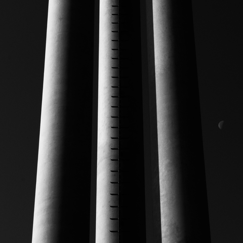A square black-and-white photograph framed with a white border, showing three massive vertical concrete posts, supporting a billboard structure. The central post features a mounted metal ladder running straight up its length, creating a rhythmic pattern of shadows. All three columns are side-lit, revealing smooth gradients and emphasizing their cylindrical shapes. In the dark sky to the right, a half-moon appears, forming a visual echo to the rounded forms of the posts. The composition is minimal and architectural, with a strong interplay of light, shadow, and repetition.