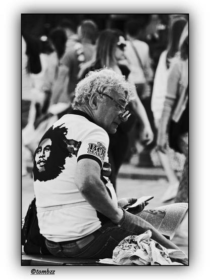 The photo shows a man sitting on a low wall. He’s holding a smartphone in his left hand, while his right hand is tucked into the pocket of his trousers. A newspaper is spread out on his lap.
The man is wearing a t-shirt with a large print of Bob Marley’s face on the back. The sleeve of the shirt bears the text “BOB MARLEY.” He has curly gray hair and is wearing dark sunglasses.
In the background, there’s a lively street scene with many people in motion, adding energy to the composition. The shot is black and white, in a street photography style, which emphasizes the raw, documentary feel of the moment. The framing captures an authentic slice of city life.