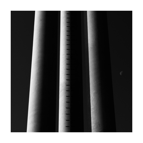 A square black-and-white photograph framed with a white border, showing three massive vertical concrete posts, supporting a billboard structure. The central post features a mounted metal ladder running straight up its length, creating a rhythmic pattern of shadows. All three columns are side-lit, revealing smooth gradients and emphasizing their cylindrical shapes. In the dark sky to the right, a half-moon appears, forming a visual echo to the rounded forms of the posts. The composition is minimal and architectural, with a strong interplay of light, shadow, and repetition.