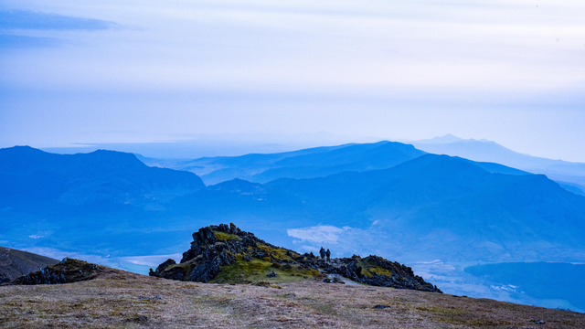 Blue mountains below with people walking off the edge of the top one in the foreground with haze in the valley and sky 