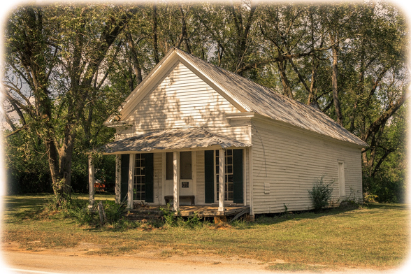 A small, weathered white wooden house with a steep gabled roof sits in a rural setting surrounded by mature trees. The single-story structure features a covered front porch supported by simple wooden posts, with French doors and windows flanked by dark shutters. The building appears to be historic, with horizontal wood siding that shows signs of age and weathering. A metal roof covers the main structure, and there's a small chimney visible. The house is set on what appears to be a raised foundation, with wooden steps leading to the porch. The surrounding yard is a mix of grass and bare earth, with large trees creating a canopy overhead. The photograph has a vintage or sepia-toned quality with soft, vignette-style edges that give it an aged appearance.