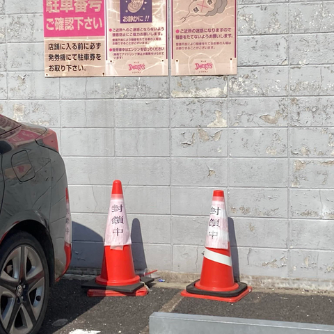 The image shows a grey car parked near a wall with peeling paint. Two traffic cones with the words "封鎖中" (meaning "closed" in Japanese) are placed on the ground. Above them, there are posters with instructions related to