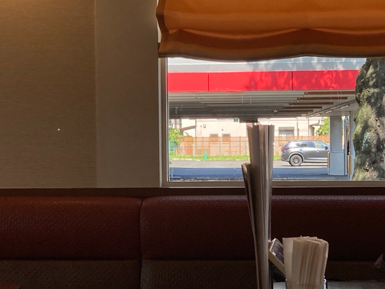 A view from inside a restaurant looking out a window. The image shows a red overhead canopy, a parking lot, and a car passing by. There are napkins and utensils on a table in the foreground.