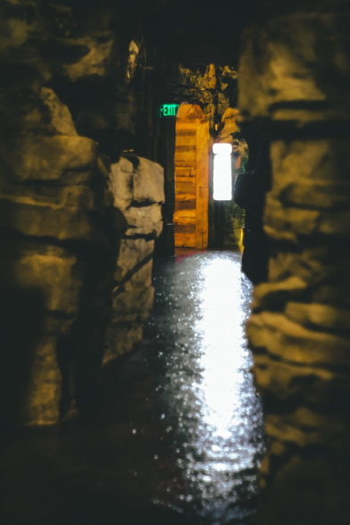 A dim, narrow stone corridor glistens with moisture under low light, leading to a warm wooden doorway glowing beneath a green exit sign, evoking a mysterious and slightly eerie atmosphere.