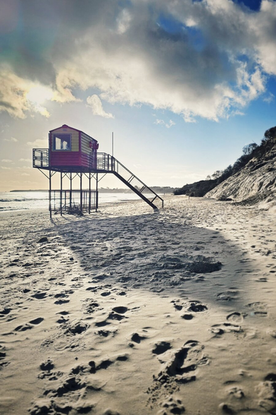 A red and yellow lifeguard hut stands elevated on stilts on a wide, sandy beach. Numerous footprints mark the sand, stretching into the foreground. In the background, a gentle slope of sand and some trees are visible under a partly cloudy sky with patches of bright sunlight. The light casts long shadows across the beach from the hut and a nearby fence.