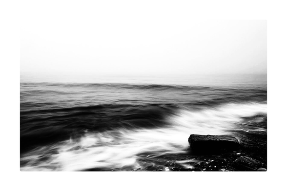 A black and white photo showing a long exposure view of the ocean touching a rocky beach in Maine.