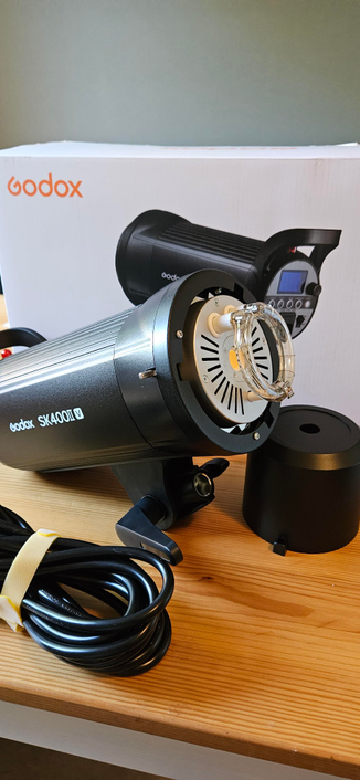 A picture of a just unpacked Godox SK400II-V studio flash light, as well as a cable and a mount protection. In the background is the huge white box it came in.