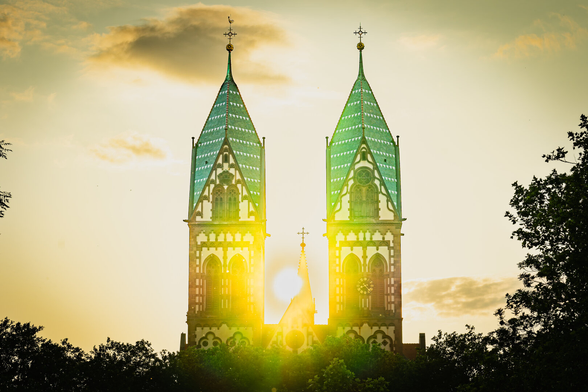A bright sunset illuminates twin church towers with green-tiled roofs and intricate architectural details. The sun sits directly between the towers, creating a glowing halo effect. Silhouetted trees frame the scene on both sides, and a partly cloudy sky is tinged with warm golden hues.