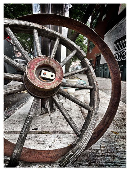 A discarded wooden wagon wheel with a detached metal rim leans against a power pole on a residential city sidewalk. The wheel’s hub has remnants of flaking red paint, and its spokes radiate out to a wooden inner rim that is broken and splintered in several places. One of the spokes is mostly missing, with only a sliver of wood attached to the hub. The detached outer metal rim is rusted red. In the background are glimpses of parked cars, trash bins lined against an apartment building, and the green foliage of trees. The image emphasizes the textures of the wooden wheel, the rusted surface of the rim, and the ridges of the sidewalk.