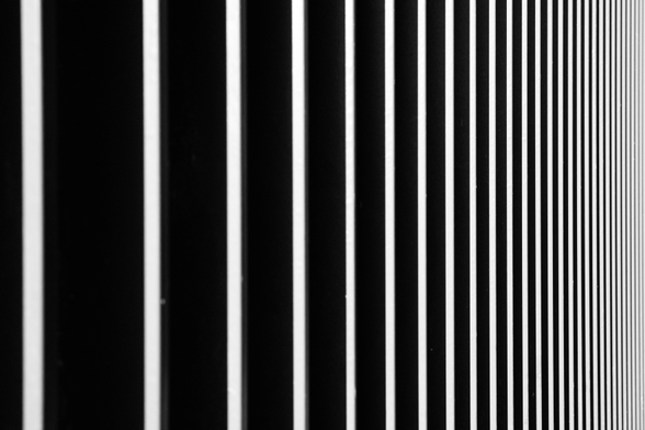 Abstract black and white picture of white vertical parallel lines, close together on the right, further apart on the left. It’s actually a metal fence caught in the evening sunlight but it looks like an art 