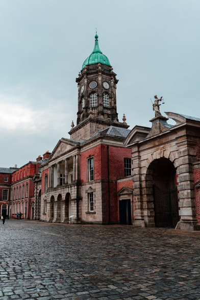 A brick road lines the way in front of an old brick building with a red facade and a tower rising from the middle.