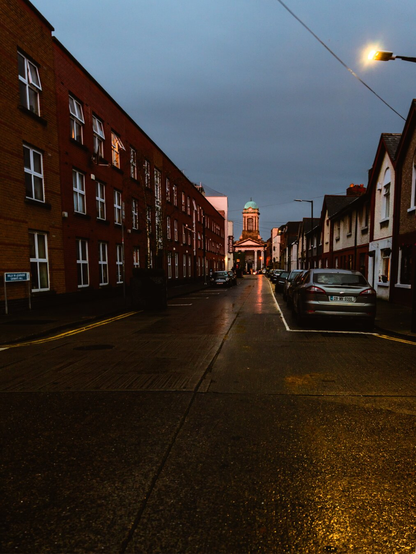 A street light illuminates the road lined with a brick apartment building leading to a building with a tower