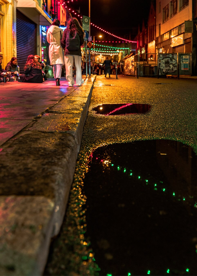 People are sitting and walking on the sidewalk at night while a puddle reflects the string lights illuminating the street at night in Dublin.