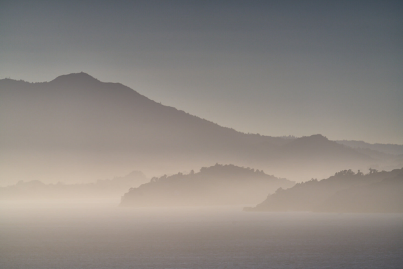 Long distance shot of Mt Tamalpais in Marin County. Angel Island and low hills are layered in the foreground. There is a haze at seal level. The photo is monochrome, but shot in full color with a long exposure and without any color adjustments.