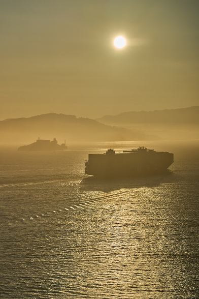 Long distance shot of Marin County with Alcatraz in the foreground and a very large and loaded MSC cargo ship crossing closer in. There is a haze at seal level. The light is golden.