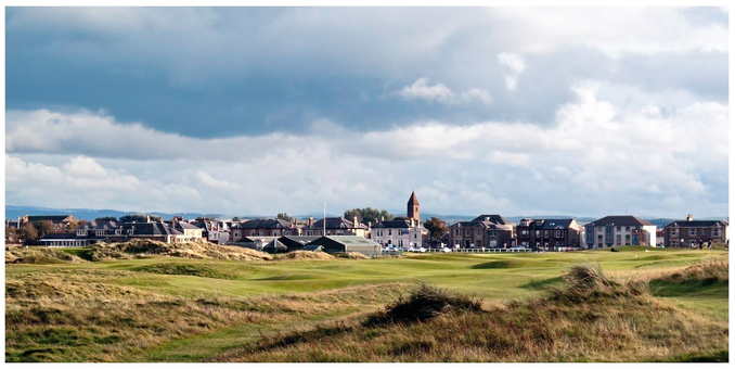 A scenic view of a golf course with rolling green hills in the foreground and a town in the background. The sky is partly cloudy, and notable buildings, including a tower, can be seen among residential structures.
