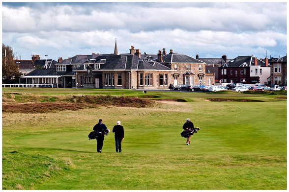 Three golfers walk across a green area towards a large building, likely a clubhouse, in the background. The sky is partly cloudy, and parked cars are visible.