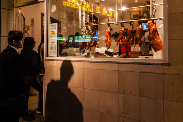 A man gazes at cooked chickens hanging on hooks in the window of a Chinese food shop at night. His shadow, bigger than him, is cast on the wall below. The shop window has blurred reflections of signs with English and Chinese writing on them. Inside the shop, a man with a Chinese chef's knife raised in his hand, is visible between the chickens.