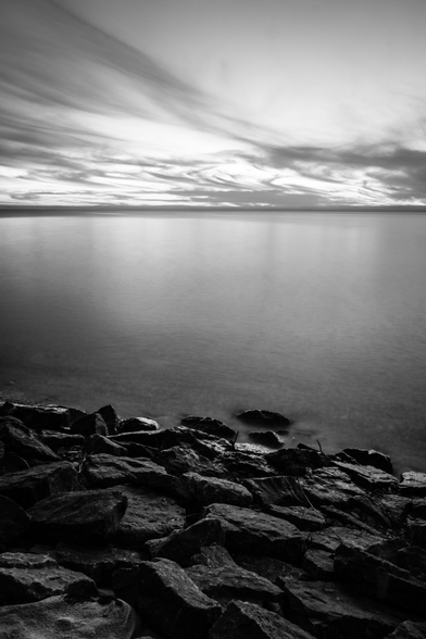The shoreline of Empire Beach with large rocks in the foreground and Lake Michigan stretching to the horizon. The sky is partly cloudy with streaks of clouds across it.