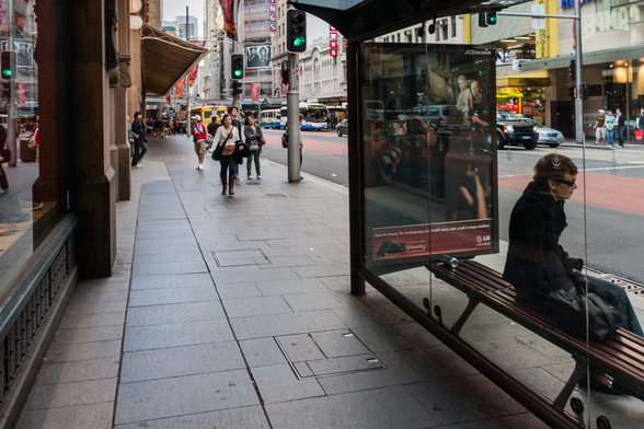 In Sydney's main Street, George Street, a lone woman sits in a bus shelter on the right. In the background people are walking along the pavement and traffic and buildings can be seen.