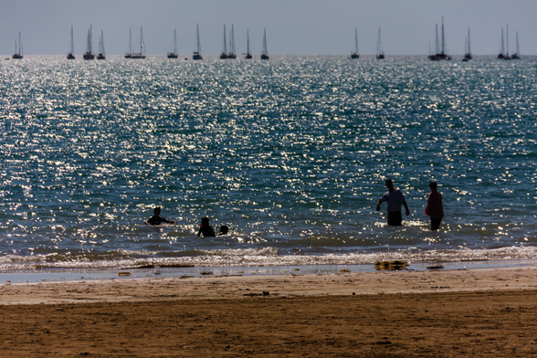 There's a small amount of beach in the foreground, and most of the rest of this image is of the ocean shining in the sunlight. Near the shore, a few bathers are in silhouette, and on the horizon there are many sailing boats.