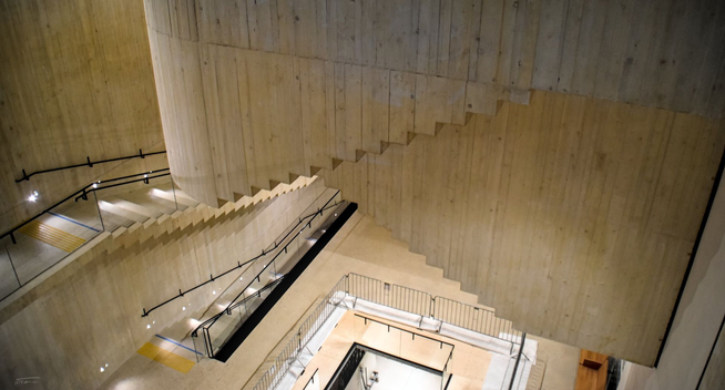 Das Foto zeigt ein faszinierendes Spiel aus Perspektiven und geometrischen Formen im Inneren des Wien Museums. Der Fokus liegt auf einem modernen Treppenhaus mit mehreren Ebenen und Richtungswechseln. Die Betonwände und -treppen dominieren die Komposition, wobei die Treppen teils parallel, teils im rechten Winkel zueinander verlaufen. Sie werfen harte Schatten, die die Illusion von Tiefe und Komplexität verstärken.

Die Architektur wirkt fast surreal – ein klarer Verweis auf den Künstler M.C. Escher, der für seine unmöglichen, perspektivisch verwirrenden Bilder bekannt ist. Die geschwungene, spiralförmige Raumstruktur und die überlappenden Treppen erzeugen eine Art visuelle Illusion, die scheinbar die Schwerkraft und Orientierung infrage stellt.

Das Licht ist gedämpft, aber akzentuiert – Spotlichter an den Wänden und entlang der Handläufe betonen die Konturen und Kanten der Architektur. Gelbe Markierungen auf den Stufen bieten nicht nur Orientierung, sondern setzen auch kleine, visuelle Kontrapunkte zur sonst warm-grauen Farbpalette des Sichtbetons.
